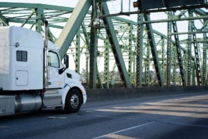 Clásico y potente semirremolque blanco de plataforma grande con compartimento para dormir en cabina grande para rutas de largo recorrido que van por el puente interestatal del río Columbia que transporta carga comercial al punto de destino